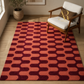 A rectangular area rug with a retro mid-century modern pattern of alternating coral orange and deep burgundy rounded-rectangle shapes, styled on a light wood floor next to a wooden armchair with a white cushion.