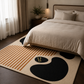 A modern area rug with abstract black shapes and tan stripes on a cream background, styled on a dark wood floor in a bedroom next to a bed with neutral bedding.