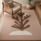 A rectangular area rug in a warm beige tone with a large stylized brown coral or tree-like motif and a white crescent shape, styled in a bright living room beside a wooden armchair and vase.
