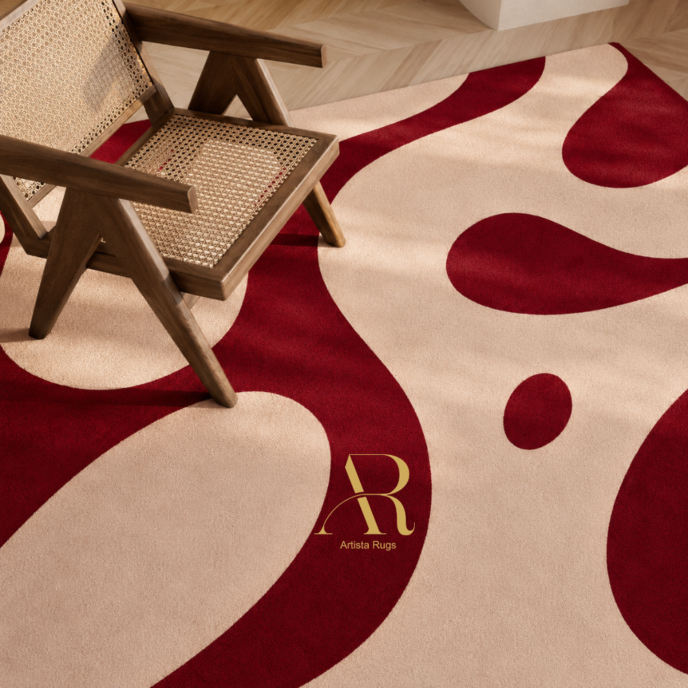 A close-up of a modern area rug with an abstract wavy pattern in deep burgundy and warm cream, styled with a wooden cane-back chair. The cream section features a gold 'AR' monogram logo and the brand name 'Artista Rugi'.