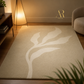 A rectangular area rug in warm beige with a large off-white botanical leaf pattern, styled in a modern living room setting with a floor lamp, wooden side table, and a neutral-colored armchair.