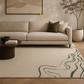 A modern living room scene featuring a large beige area rug with dark green wavy line patterns, styled with a light beige sofa, dark olive and cream pillows, a round dark wood coffee table, and a potted tree in the background.