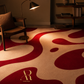 A modern area rug with an abstract wavy pattern in deep red and cream, styled in a warm, dimly lit living room with a wooden armchair, floor lamp, and bookshelf.