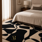 A modern abstract area rug in black and beige with a gold 'R' logo in the corner, styled in a bedroom setting under a bed.