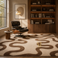 A large area rug with a wavy, organic brown pattern on a cream background, styled in a modern, warm-toned living room with a black leather lounge chair, wooden bookshelves, and a floor lamp.