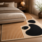 A modern area rug styled in a bedroom setting, featuring a cream base with rust-orange vertical stripes, large black abstract shapes, and a small gold monogram 'R'.