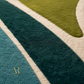Close-up of a plush area rug with a modern abstract pattern in dark teal, forest green, olive yellow, and cream, featuring a small gold monogram logo in the lower left corner.