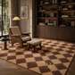 A large checkered area rug in warm beige and dark brown tones, styled in a cozy, sunlit living room with a mid-century modern armchair, wooden bookshelves, and a matching ottoman.