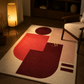 A modern abstract area rug with bold geometric shapes in red, burgundy, and beige, styled in a warm, dimly lit living room next to a wooden chair and a glowing floor lamp.