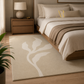 A beige area rug with a large white leaf motif, styled in a modern bedroom next to a wooden bed and nightstand.