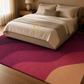 A large area rug with a wavy abstract pattern in magenta, burgundy, and tan, shown in a styled bedroom setting under a bed with neutral bedding.