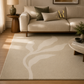 A large beige area rug with a large-scale abstract leaf pattern in cream, styled in a warm, modern living room with a beige sofa, wooden side table, and soft natural light.
