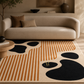 A modern abstract area rug with black organic shapes and warm orange stripes on a cream background, styled in a living room setting under a beige sofa.