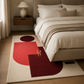 A modern geometric area rug in cream, red, and burgundy, styled on a light wood floor in front of a bed with neutral bedding and a bedside table.