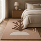 A rectangular area rug in a warm taupe-brown color, styled in a modern bedroom setting. The rug features a large dark brown coral-like silhouette and a cream-colored semi-circle at the bottom, with a gold 'R Aubrey Riley' logo in the bottom right corner.