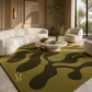 A modern living room scene featuring a large olive green area rug with dark olive abstract wavy patterns. The room is styled with a curved white sofa, a marble-top coffee table, two cream armchairs, and warm beige walls with decorative panels.