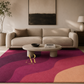 A modern living room scene featuring a large area rug with wavy abstract patterns in magenta, burgundy, and peach. The room is styled with a beige sofa, a round stone coffee table, and a gold 'Artsila Rugs' logo printed on the rug.