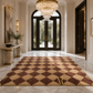 A large rectangular area rug with a brown and beige diamond pattern is displayed in a luxurious entryway with marble floors, a crystal chandelier, and dark wood doors.
