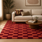 A modern living room scene featuring a large area rug with a bold geometric pattern in terracotta orange and deep burgundy. The rug is styled on a light wood floor in front of a cream-colored sofa with decorative pillows, a round wooden coffee table, and a potted plant in the background.