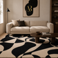 A modern living room scene featuring a large abstract black and beige patterned area rug under a cream-colored sofa, with a dark wood coffee table and wall art in the background.