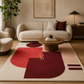 A modern living room featuring a large area rug with abstract geometric shapes in cream, pink, red, and burgundy, styled with a beige sofa, wooden coffee table, and potted plant.