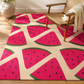 A rectangular area rug with a bright pink watermelon slice pattern on a cream background, styled in a room with a wooden chair and a potted plant.