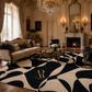 A large area rug with an abstract black and cream wavy pattern is shown in a luxurious, ornate room with a fireplace, chandeliers, and antique furniture. The rug features a gold 'Arts & Riches' monogram logo in the lower center.