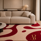 A modern beige fabric sofa in a styled living room, placed on a large area rug with bold abstract red and beige wavy patterns. A round marble-top coffee table is in the foreground, with a wall lamp, curtains, and framed wall art in the background.