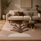 A beige living room scene featuring a large area rug with a dark brown abstract tree design and a cream semicircle at the bottom. A light beige sofa, round wooden coffee table, potted plant, and warm ambient lighting are arranged around the rug.