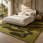 A modern bedroom scene featuring a large olive green area rug with abstract dark green wavy patterns and a gold 'Riviera' logo. The rug is styled under a cream upholstered bed with white linens, flanked by a wooden nightstand and a large window looking out onto green trees.