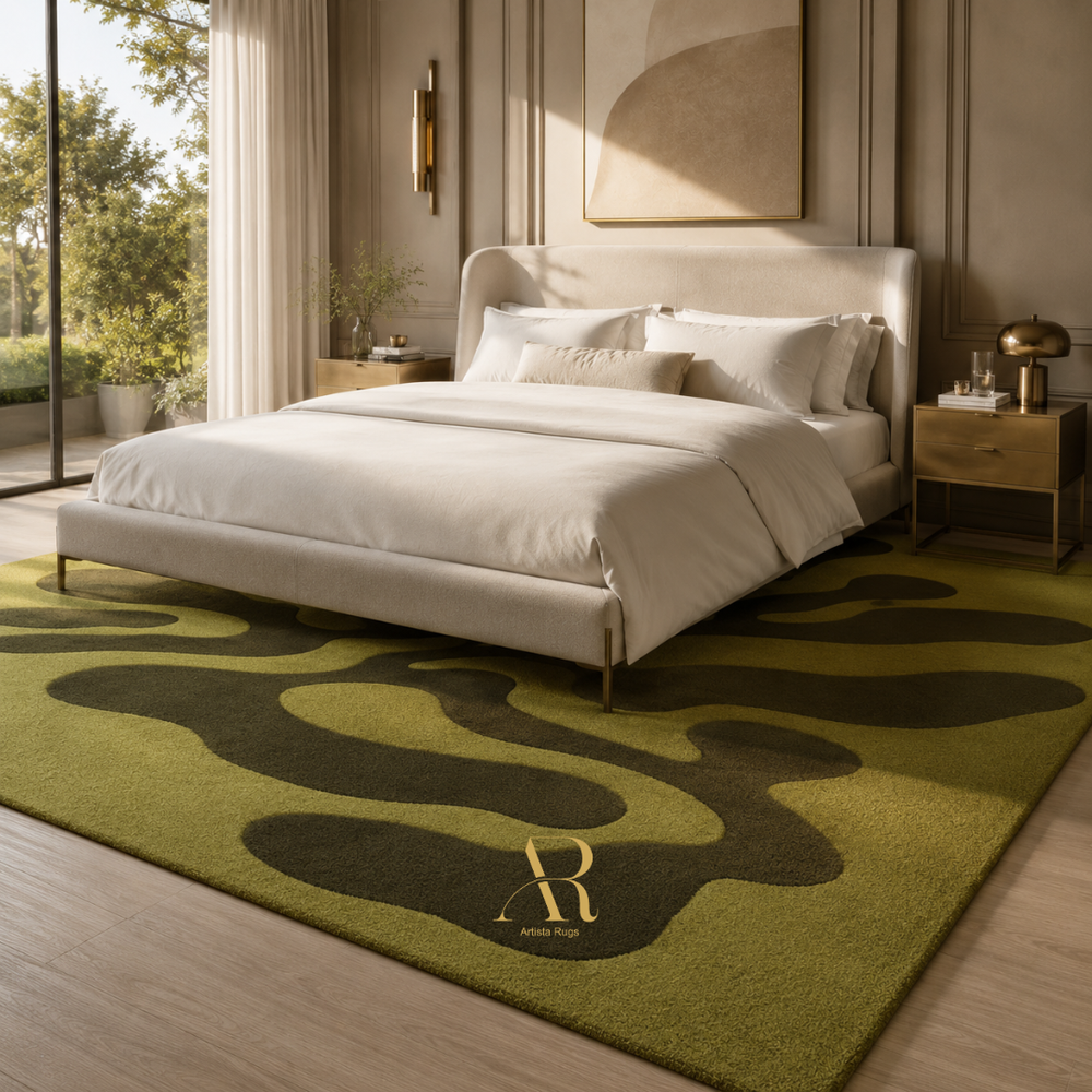 A modern bedroom scene featuring a large olive green area rug with abstract dark green wavy patterns and a gold 'Riviera' logo. The rug is styled under a cream upholstered bed with white linens, flanked by a wooden nightstand and a large window looking out onto green trees.