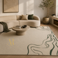 A large beige area rug with a dark green wavy line pattern and a gold monogram logo, styled in a modern living room setting with a curved beige sofa and a round stone coffee table.