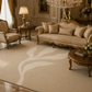 A large beige area rug with a large white leaf motif, styled in a luxurious, ornate gold and beige living room with a matching sofa, armchair, and chandelier.