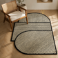 Three interlocking modern rugs with a black and beige striped pattern, styled on a light wood floor next to a wooden armchair.