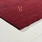 A close-up of a deep burgundy red area rug with a textured, plush pile and a small gold 'R' monogram logo, shown on a light-colored tiled floor.