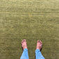 An olive green textured area rug with a small gold monogram logo in the lower right corner, displayed flat against a white background.