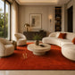 A luxury modern living room featuring a large rust-red area rug with a gold monogram logo, styled with cream-colored curved sofas, a round stone coffee table, and warm ambient lighting.