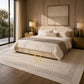 A styled bedroom scene featuring a large cream-colored area rug with a black dotted border and a gold monogram logo in the center. The rug is laid out under a bed with neutral beige and white bedding, flanked by wooden nightstands and lamps, with a large window showing an outdoor view in the background.