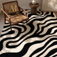 A modern black and cream wavy abstract patterned area rug styled in a room setting with a wooden cane armchair and a small wooden side table.