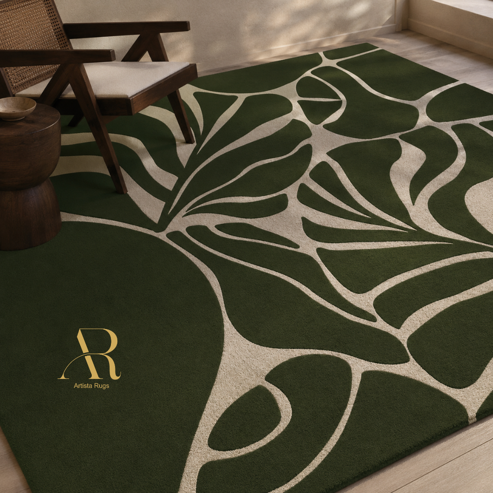 A dark green area rug with a large beige leaf-like abstract pattern, styled in a modern room setting with a wooden chair and side table. The brand logo 'Artista Rugs' is visible in the bottom-left corner.