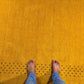 A top-down view of bare feet standing on a textured mustard yellow rug with a pattern of small black dots along the edge, paired with blue jeans.