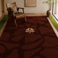 A large rectangular area rug in dark brown with a tonal abstract organic pattern and a central beige shell motif, styled in a modern living room setting with a wooden armchair, green grass-like strips, and a vase.