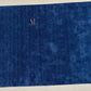 A close-up of a deep blue textured fabric with a small gold embroidered monogram logo reading 'R' near the upper left area, shown against a light neutral background.