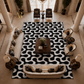 A large rectangular area rug with a bold black and white abstract wavy pattern, styled in a grand, high-ceilinged classical living room with cream sofas, dark wood chairs, and marble columns.