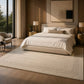A large cream-colored area rug with a subtle geometric border pattern, styled under a bed in a modern, warm-toned bedroom with wood paneling and natural light.