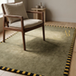 Wooden armchair with beige cushions on a green rug with yellow and black border in a room.