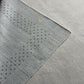A close-up of a light gray textured fabric with a diagonal dotted pattern and a small gold monogram logo, laid over a neutral gray background.