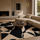 A modern living room scene featuring a large area rug with a bold black and beige abstract organic pattern. The rug is styled in a contemporary interior with a beige sectional sofa, a round wooden coffee table, a fireplace, and a large window.
