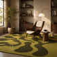 A modern living room scene featuring a prominent area rug with an organic wavy pattern in olive green and dark brown. The room is styled with a mid-century modern wooden armchair, a small wooden side table, a floor lamp, and a potted bamboo plant.