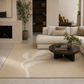 A beige area rug with a large white abstract leaf pattern, styled in a modern living room with a cream sectional sofa, a dark wood coffee table, and a fireplace.